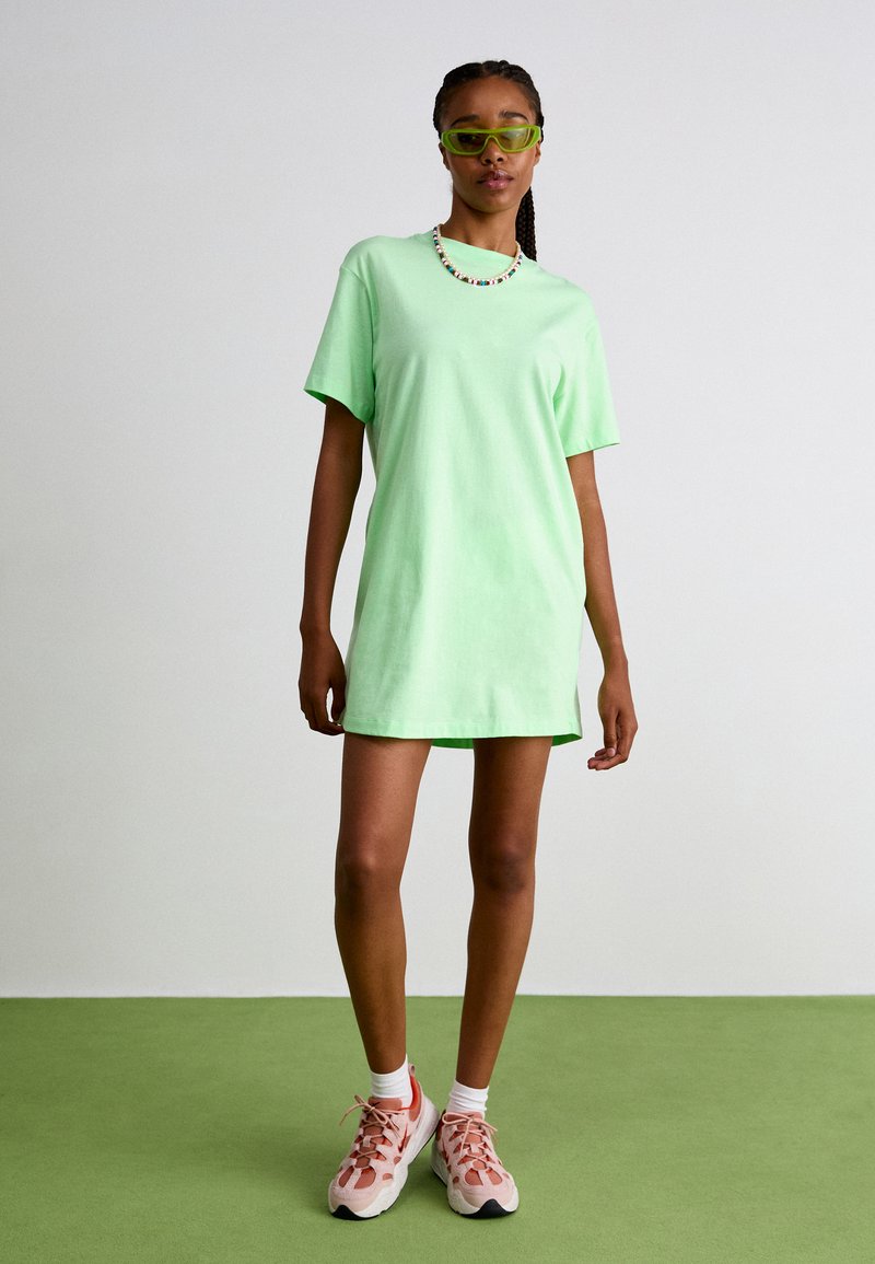 Vestido corto de algodón verde con mangas cortas, de corte relajado, combinado con zapatillas rosa claro, realzado con gafas de sol verdes y un collar de cuentas.