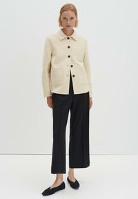 Giacca color crema in misto lana con quattro tasche, bottoni neri e colletto, abbinata a pantaloni neri a gamba larga e scarpe nere basse.