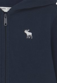 Granatowa bluza na zamek wykonana z miękkiej tkaniny, z małym białym haftowanym emblemą łosia na lewej stronie klatki piersiowej.