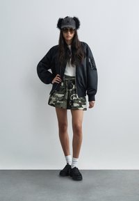 Μαύρο μπουφάν bomber, λευκό πουκάμισο, φούστα καμουφλάζ, μαύρο καπέλο με τεχνητές γούνας, γυαλιά ηλίου, λευκές κάλτσες, μαύρα παπούτσια χωρίς κορδόνια με σχέδιο πλέγματος.