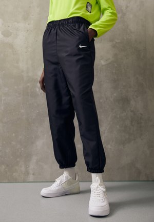 Persoon draagt zwarte Nike joggers met elastische boorden, witte sneakers, witte sokken en een neongroen longsleeve tegen een grijze, getextureerde muur.