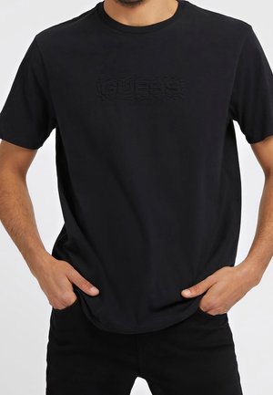 Man draagt een zwarte Guess T-shirt met reliëflogo, handen in de zakken, getoond van nek tot heupen tegen een witte achtergrond.
