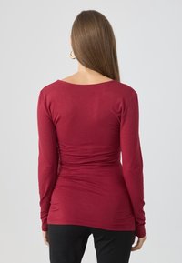 Kobieta z długimi prostymi włosami, ubrana w dopasowany czerwony top z długim rękawem i czarne spodnie, stojąca tyłem na jednolitym jasnym tle.