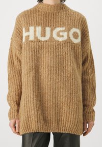 Pull en maille marron à col montant, arborant le logo blanc "HUGO" sur la poitrine. Coupe ample avec des poignets et un ourlet côtelés.