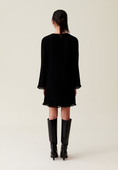 Robe noire plissée avec manches longues et ourlet volanté, associée à des bottes noires jusqu'aux genoux avec un nez pointu.