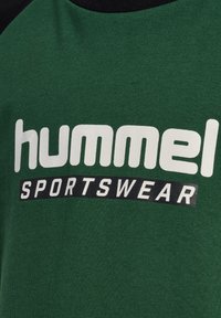 Grünes Baumwoll-T-Shirt mit schwarzen Akzenten, mit großem weißen Text "hummel" und "SPORTSWEAR" auf der Vorderseite in moderner Schrift gedruckt.
