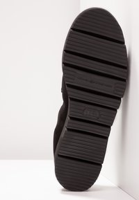 Semelle extérieure en caoutchouc noir avec texture rainurée, arborant le logo de la marque. Le côté de la chaussure présente une tige en tissu foncé avec une finition lisse et un design minimaliste.