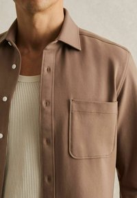 Cămașă băiețesc de culoare marron deschis, cu guler în vârf, buzunar frontal și material texturat, purtată peste un tricou crem cu dungi.