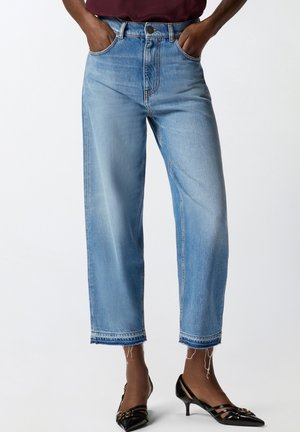 Oseba, ki nosi svetlo modre hlače do polovice meč s strganimi robovi in črne lakaste čevlje z nizkim petkom, z rokami v žepih, prikazana od pasu do nog.