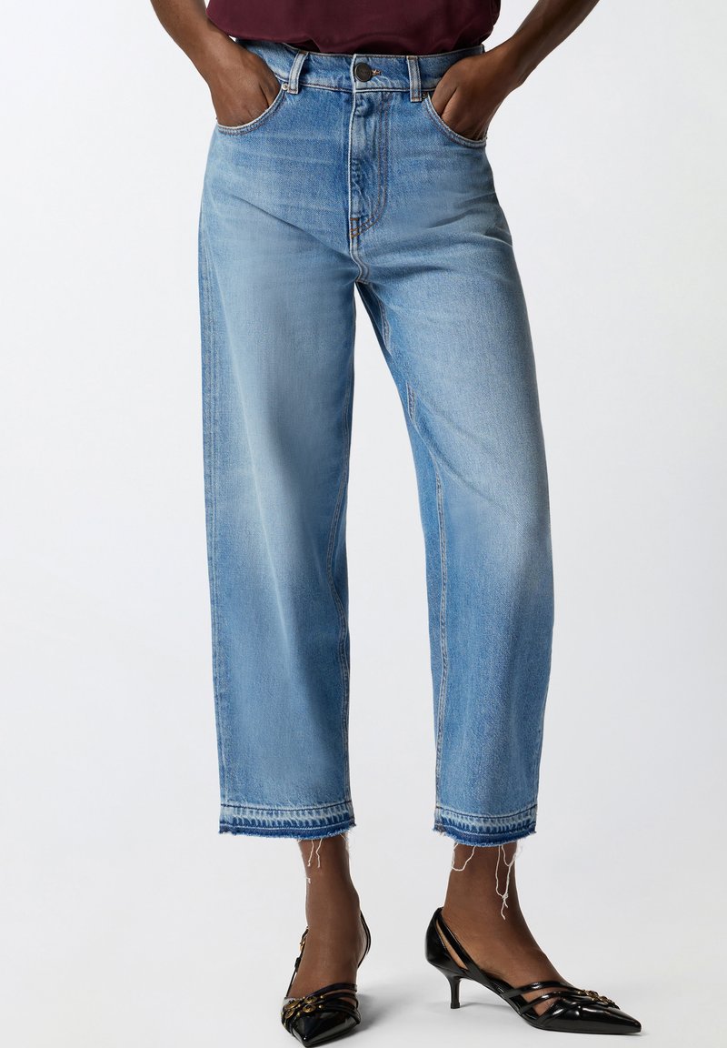 Άτομο που φορά ανοιχτού μπλε cropped τζιν με σκισμένα τελειώματα και μαύρα παπούτσια με μικρό τακούνι στιλ πατέντας, με τα χέρια στις τσέπες, εμφανίζεται από τη μέση μέχρι τα πόδια.