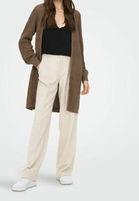 Cardigan a maglia marrone sopra una maglietta nera, abbinato a pantaloni larghi crema e sneakers bianche, che mettono in risalto una vestibilità rilassata e una superficie liscia.