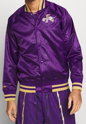 Man draagt een glanzend paars sportjack en bijpassende broek met gouden en zwarte bliksemschichtstrepen en een dinosaurus basketballogo op de borst.
