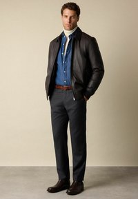 Mężczyzna w ciemnej skórzanej kurtce, niebieskiej koszuli dżinsowej, białym golfie, ciemnych spodniach, brązowym pasku i brązowych skórzanych butach na jednolitym tle.