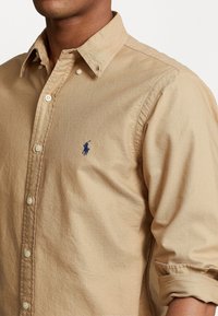 Camisa beige abotonada hecha de algodón texturizado, con cuello, tapeta abotonada y un pequeño logo azul en el pecho.