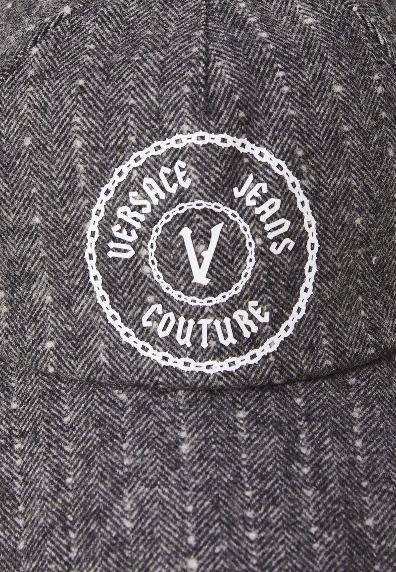 Gorra gris en patrón de espiga con un logotipo circular blanco que dice "Versace Jeans Couture" y una destacada "V" en el centro. Material de mezcla de lana.