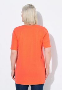 Krátký rukáv, oranžová tunika s kulatým výstřihem, vyrobená z měkké látky, s mírně delším lemem a hladkou strukturou.
