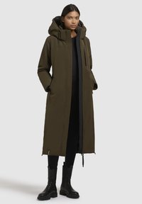 Olivgrön lång parka med huva. Har dragkedja och tryckknappar, sidofickor och rak passform. Tillverkad av lättviktsmaterial.