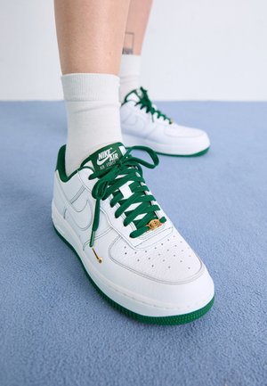 White Nike Air Force sneakers with green soles and laces worn with white socks on a light blue carpeted floor.