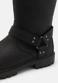 Bota de cuero texturizado negro con una correa y detalles de metal, que presenta una suela resistente y un acabado suave. Diseñada para la durabilidad.