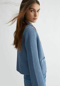 Cardigan azzurro chiaro con texture, dal taglio ampio, rifinito con bottoni e un motivo intrecciato, abbinato a pantaloni blu coordinati.