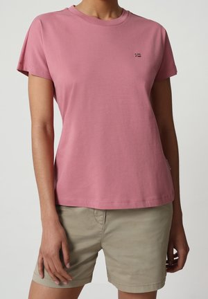 Vrouw draagt een effen stofroze T-shirt met korte mouwen en beige knielange korte broek tegen een effen achtergrond.