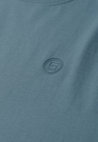 Gros plan sur un tissu bleu avec un petit logo circulaire en relief comportant une lettre stylisée au centre près de l'encolure.