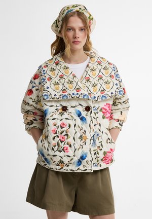 Vrouw draagt een bloemen gewatteerd jack en bijpassende hoofddoek, gecombineerd met olijfgroene shorts, staand met handen in de zakken.