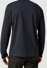 Hombre con una camisa de manga larga lisa de color azul marino oscuro y pantalones oscuros, de pie con la espalda hacia la cámara frente a un fondo liso.