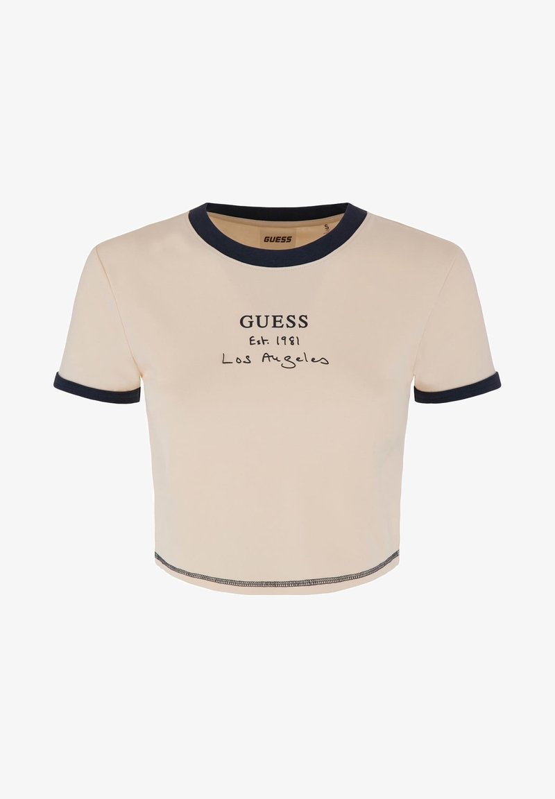 T-shirt beige à manches courtes et crop, avec un bord bleu marine. Il présente un texte imprimé "GUESS Est. 1981 Los Angeles" sur le devant. Texture lisse.