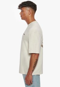 Hellbeige T-Shirt aus Baumwolle mit kurzen Ärmeln und Rundhalsausschnitt, ausgestattet mit einem Grafikdruck auf der Rückseite und einem bestickten Logo auf der Vorderseite.