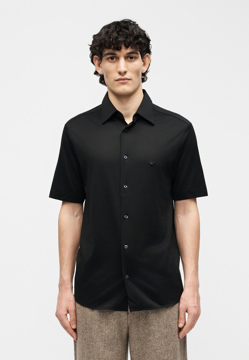 Νέος άνδρας με σγουρά μαλλιά, ντυμένος με μαύρο πουκάμισο με κοντά μανίκια και μπεζ παντελόνι, στέκεται μπροστά σε ένα απλό φωτεινό φόντο.