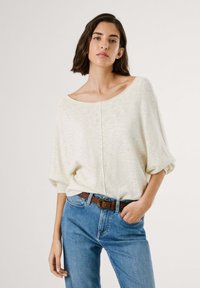 Maglione bianco oversize lavorato a maglia con scollatura ampia e maniche arrotolate, abbinato a jeans blu a vestibilità rilassata e una cintura marrone con fibbia decorativa.