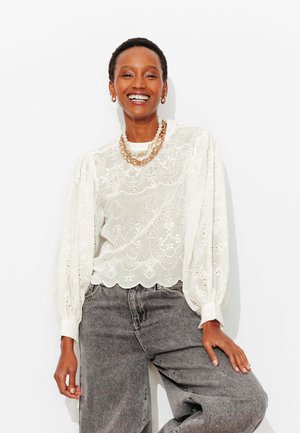 Femme souriante portant un chemisier blanc brodé à manches bouffantes, des colliers superposés en or et perles, et un jean gris taille haute.