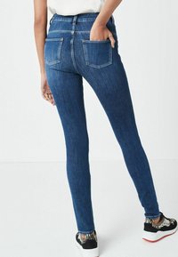 Jeans ajustados de color azul oscuro con una cinturilla alta, que cuentan con dos bolsillos traseros y una textura de denim suave. Llevados con zapatos en blanco y negro.