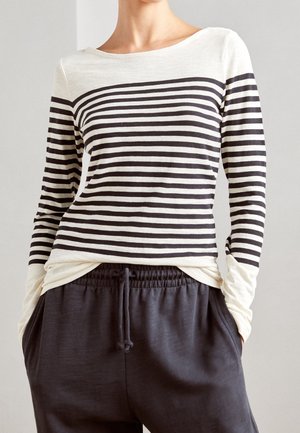 Femme portant un haut à manches longues rayé horizontalement noir et blanc, légèrement rentré dans un pantalon noir à cordons, les mains dans les poches.
