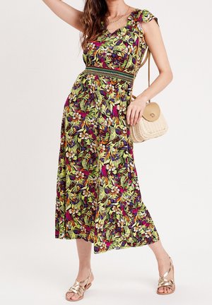Femme portant une robe midi fleurie colorée avec des manches courtes, tenant un sac bandoulière beige tissé et portant des sandales dorées à brides.