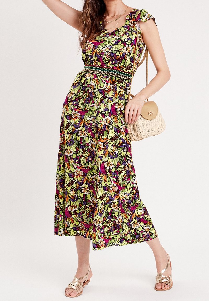Femme portant une robe midi fleurie colorée avec des manches courtes, tenant un sac bandoulière beige tissé et portant des sandales dorées à brides.