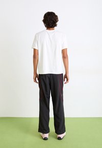 Witte katoenen t-shirt, ontspannen pasvorm, gecombineerd met zwarte broeken met rode zijkanten, en roze schoenen met een zwarte zool. Achtergrond is vlak.