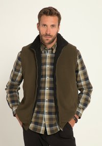 Olivgrüner ärmelloser Weste mit einem schwarzen Fleece-Kragen, getragen über einem karierten Hemd in Grüntönen, Blau und Gelb. Verfügt über Seitentaschen.