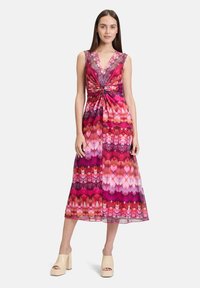 Ärmelloses Maxikleid mit tiefem V-Ausschnitt, das ein buntes Herz- und geometrisches Muster in verschiedenen Pink- und Rottönen zeigt. Knoten-Detail in der Taille.