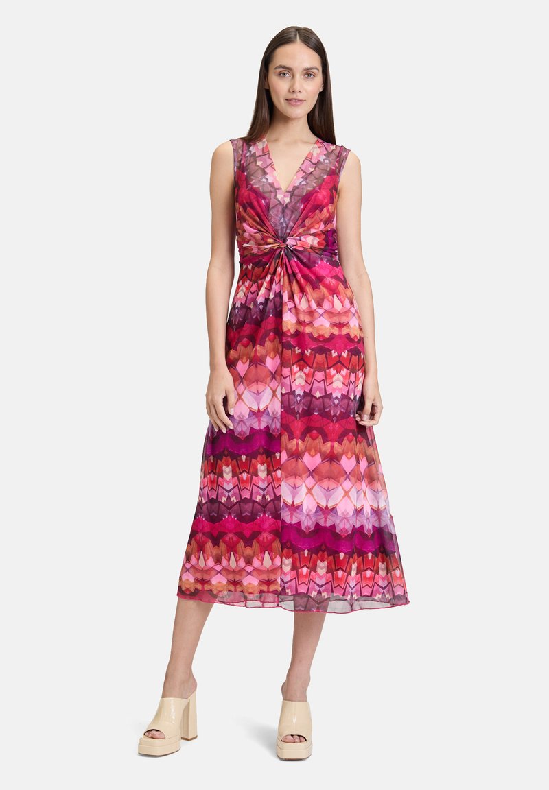 Ärmelloses Maxikleid mit tiefem V-Ausschnitt, das ein buntes Herz- und geometrisches Muster in verschiedenen Pink- und Rottönen zeigt. Knoten-Detail in der Taille.