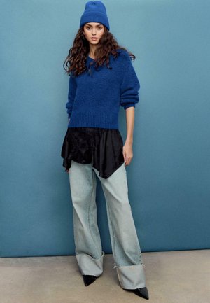 Modrý pletený sveter s pustenými ramenami, vrstvený cez čierny saténový top. Svetlomodré džíny s širokými nohavicami a oťaženými lemami, v kombinácii s čiernymi lodičkami.