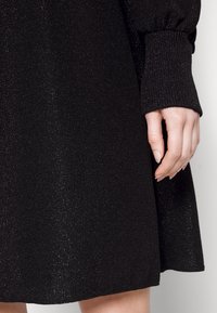 Robe noire côtelée avec un léger éclat. Comprend des manches longues texturées avec des poignets froncés et un ourlet évasé. Gros plan sur une main posée sur le tissu.