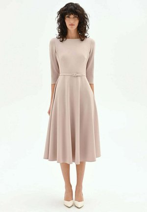 Donna con capelli scuri e ricci che indossa un abito midi beige chiaro con cintura, maniche a tre quarti e tacchi bianchi a punta, in piedi ferma.