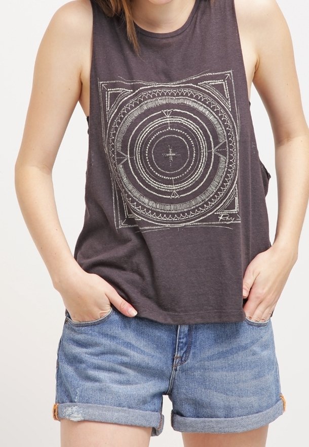 Femme portant un haut sans manches gris foncé avec un motif géométrique circulaire et un short en jean bleu retroussé, les mains dans les poches.