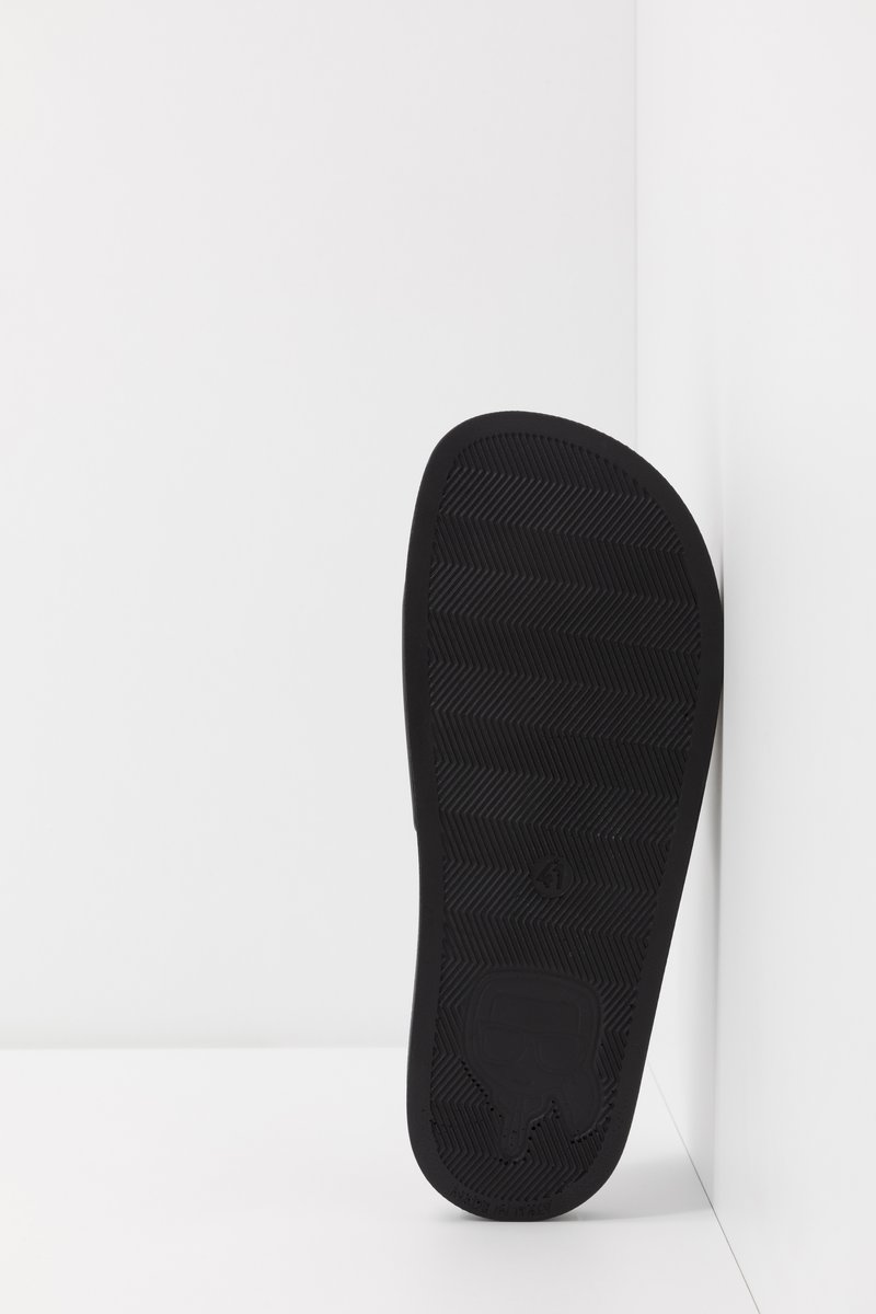 Semelle en caoutchouc noir avec un motif en chevrons, présentant un logo et des bords lisses, positionnée sur un fond blanc.