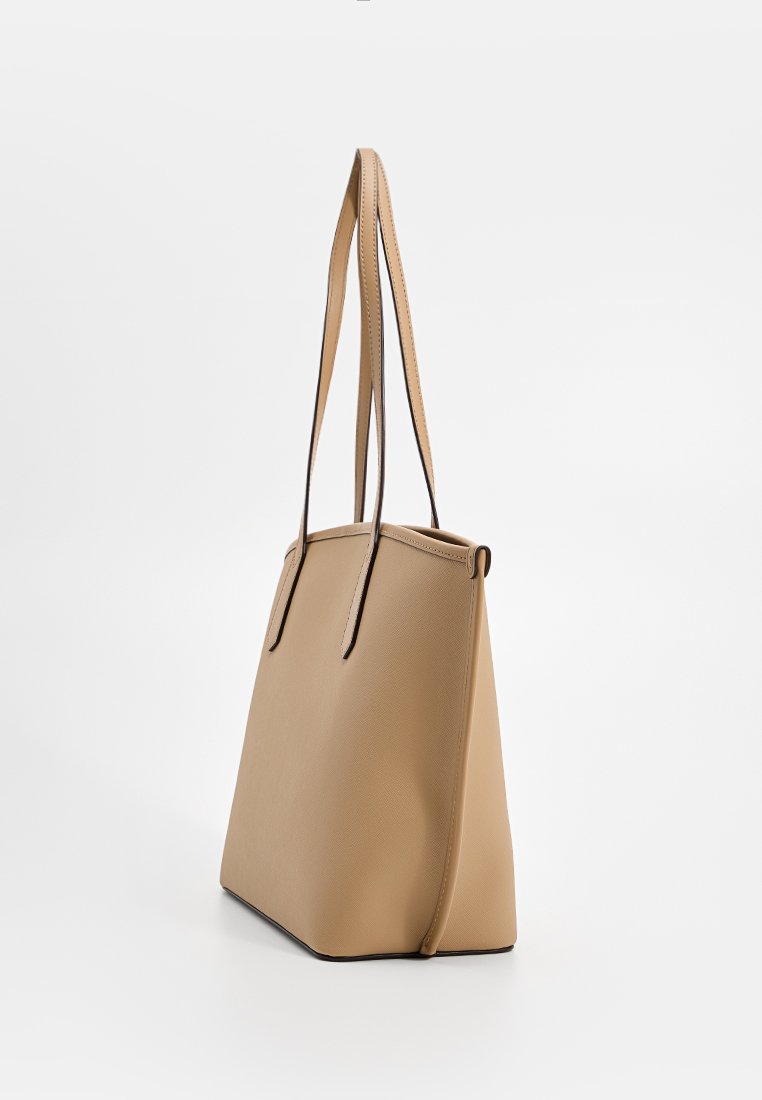 Borsa tote di forma trapezoidale beige con manici lunghi e sottili e superficie texturizzata, in posizione verticale su sfondo bianco.