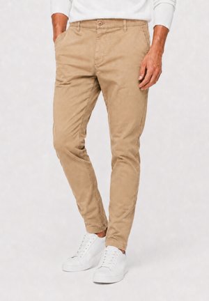 Man draagt beige slim-fit chinos en witte vetersneakers, staat met één hand in zijn zak en één hand ontspannen.