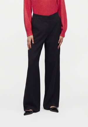 Femme portant un pantalon noir à jambes larges, des chaussures noires pointues et un haut rouge à manches longues, debout devant un fond blanc uni.