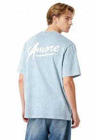 Ljusblå bomulls-t-shirt med avslappnad passform. Har vit kursiv text "Amore" på ryggen. Korta ärmar och rund halsringning.